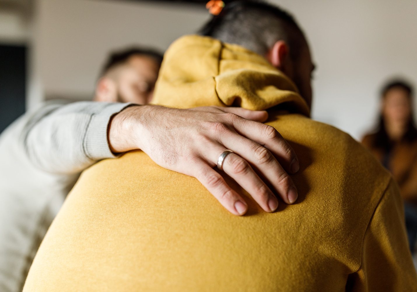 Close up shot of compassionate man embracing his friend who is sharing a heavy story regarding his mental health during a group therapy session.